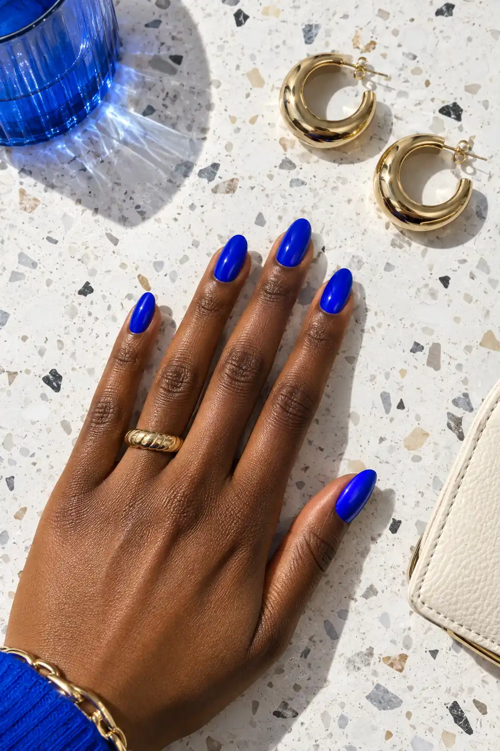 Electric cobalt blue nails on deep skin in a flat lay with gold hoop earrings